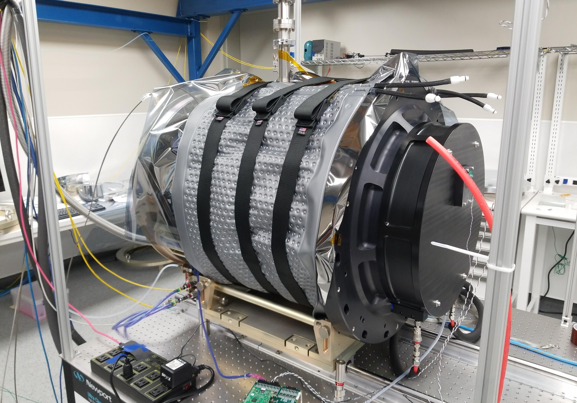 A large cylindrical scientific instrument or telescope component with reflective silver and black surfaces, mounted on a support frame in a laboratory setting with cables and equipment visible. Around the unit is a custom cooling pad provided by Polar Products that circulates cold water.