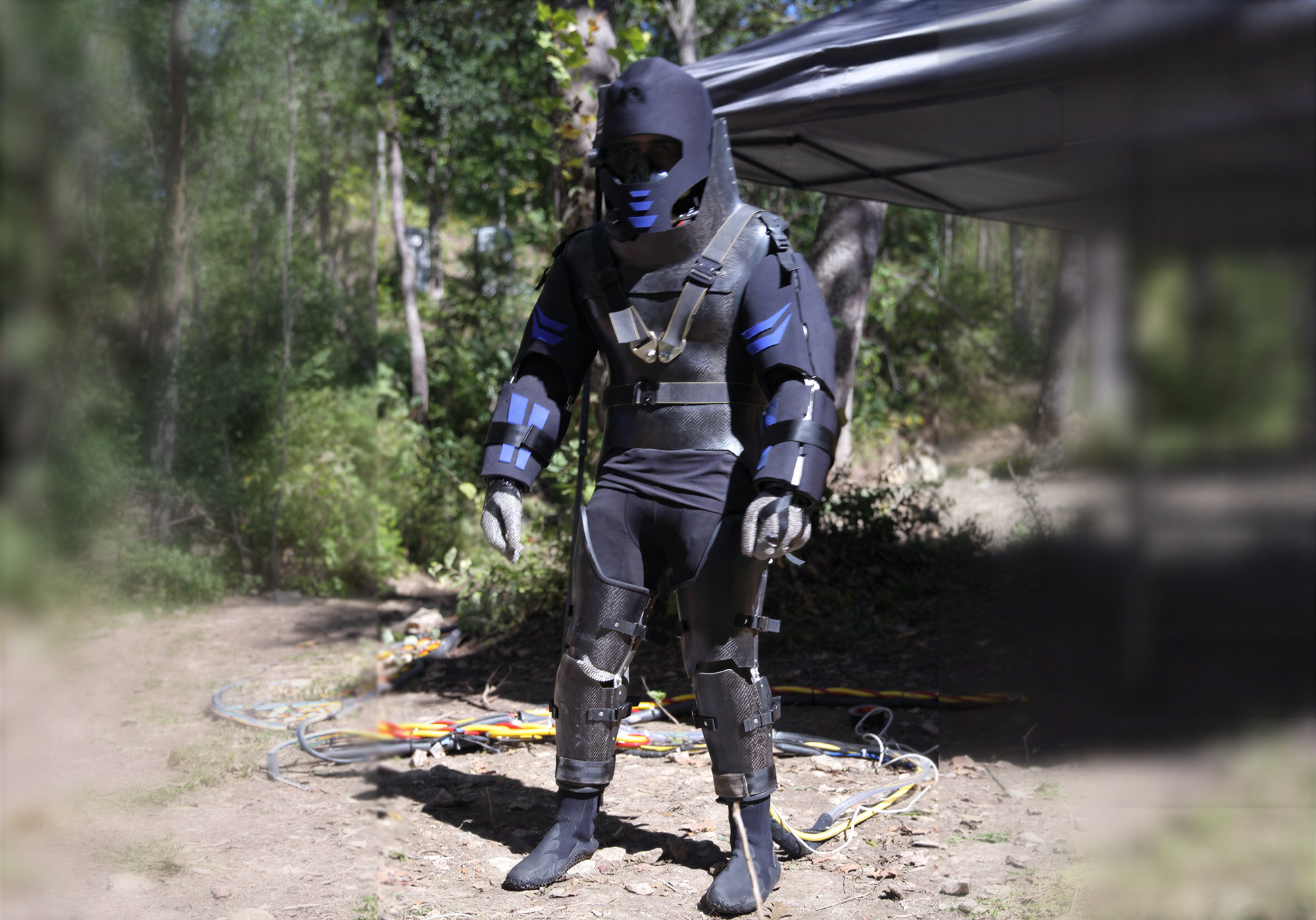 A person wearing a full-body black suit with blue accents stands outdoors on a dirt path under a canopy, with trees and equipment visible in the background. Underneath the suit is a custom body cooling device provided by Polar Products.