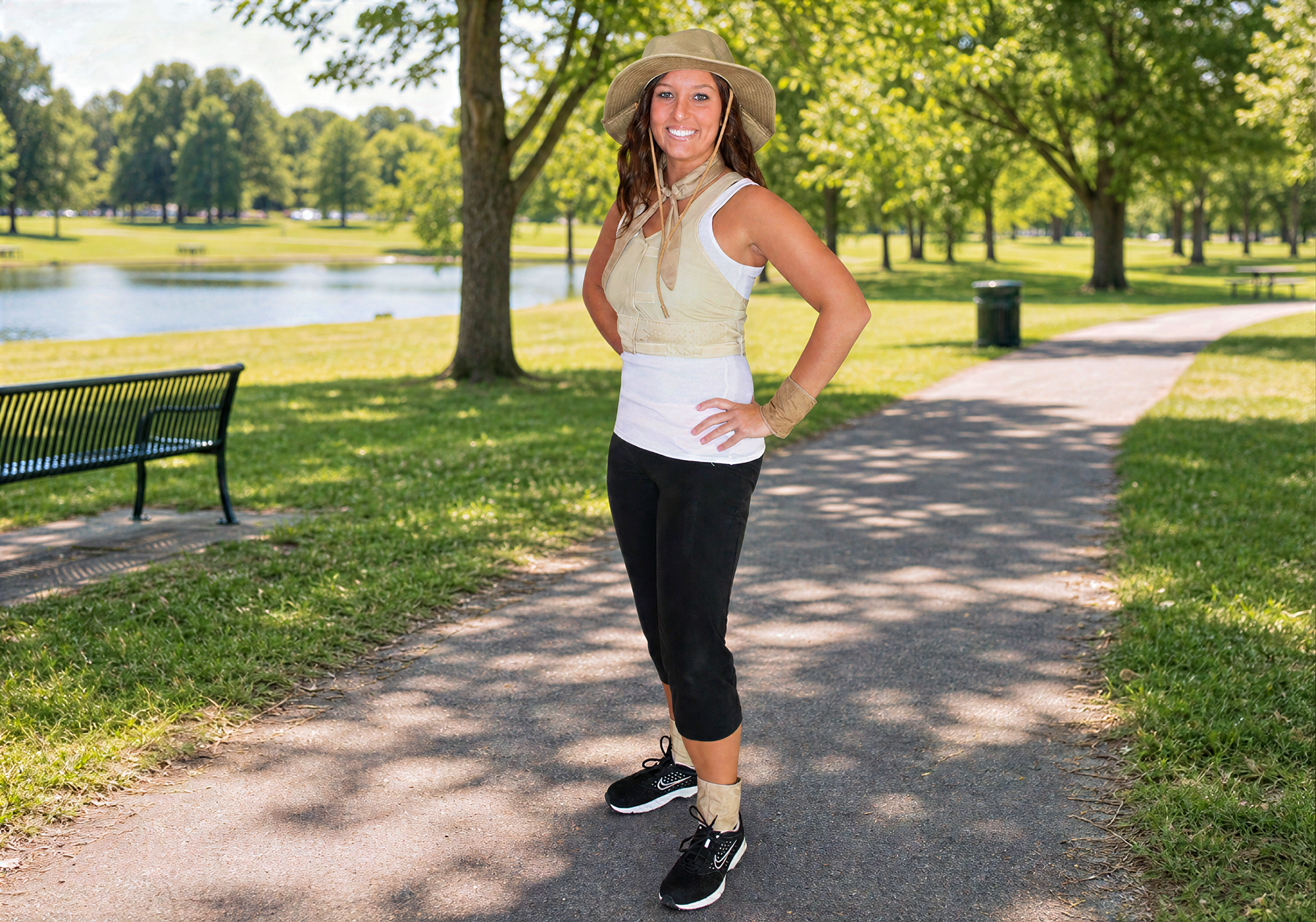 Woman standing on a path in a park with trees and a lake in the background wear various body cooling accessories from cooling ankle wraps, wrist wraps, hidden vest, neck wrap, and a bucket hat all in the khaki color.