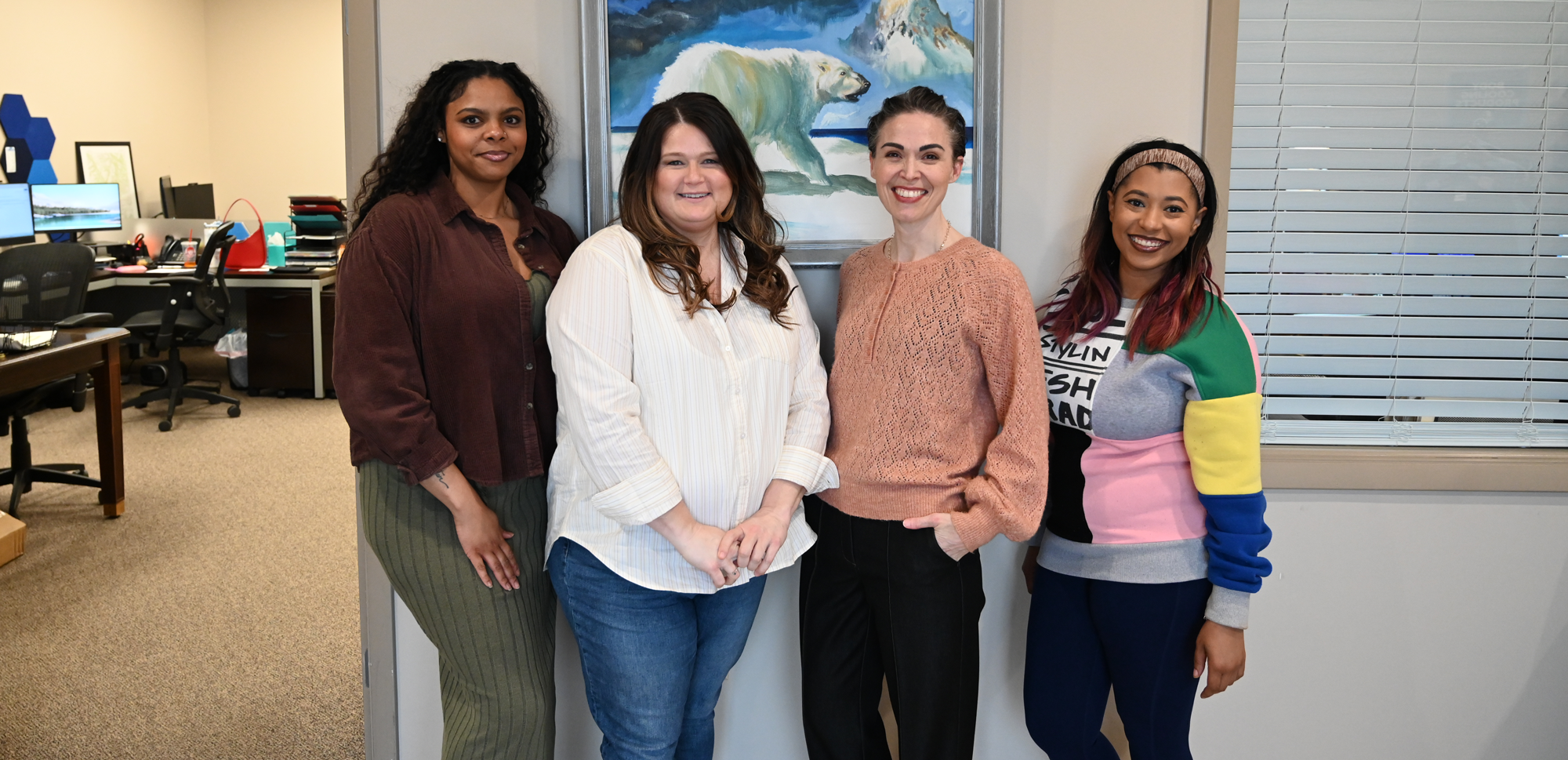 Four women standing together in an office environment, with a framed polar bear painting on the wall behind them.