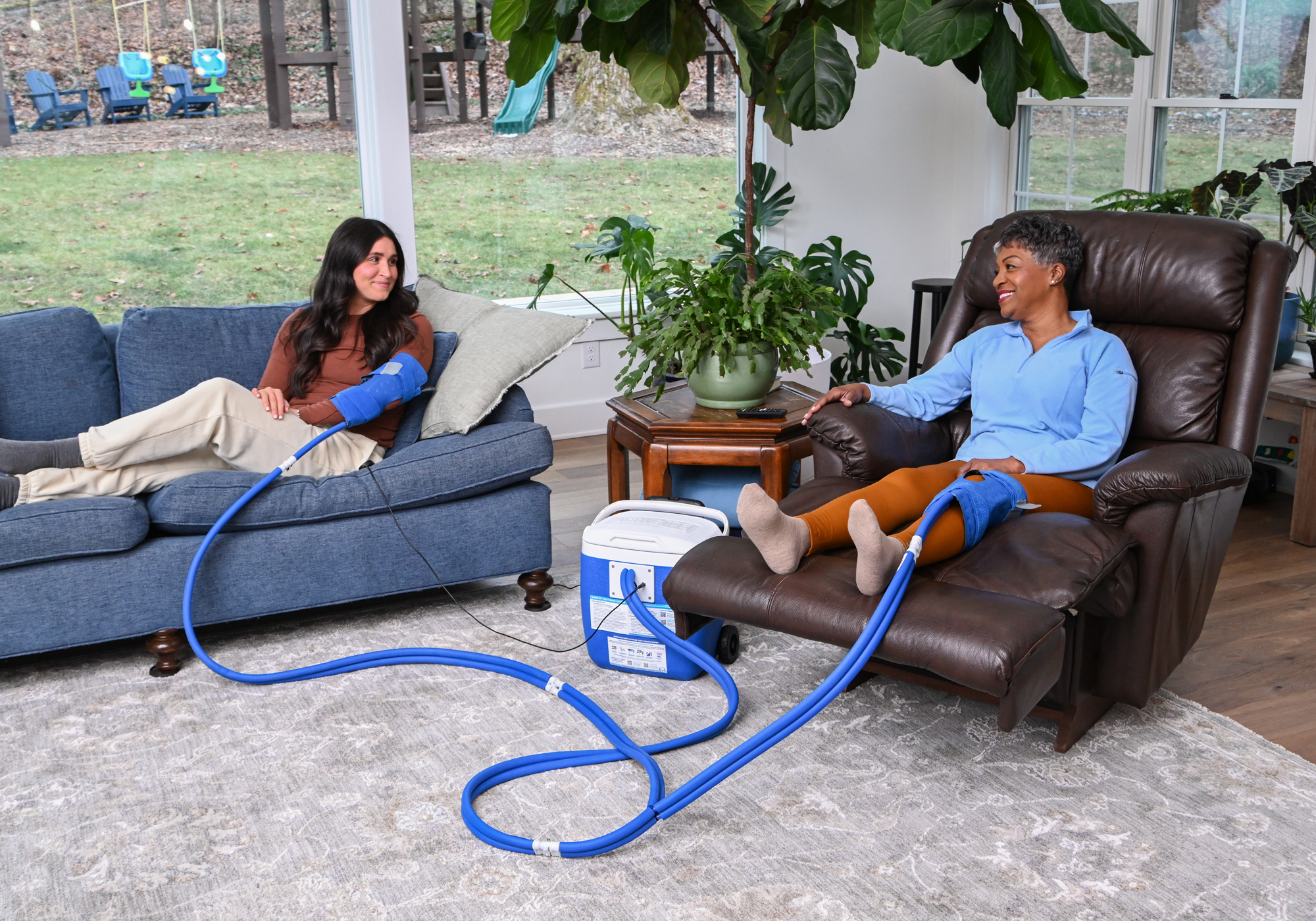Two people sitting in a living room both using an Active Ice® Knee & Joint cold therapy system in a 16-quart cooling reservoir. One woman is using it on her elbow, while the other is using it on her knee. 