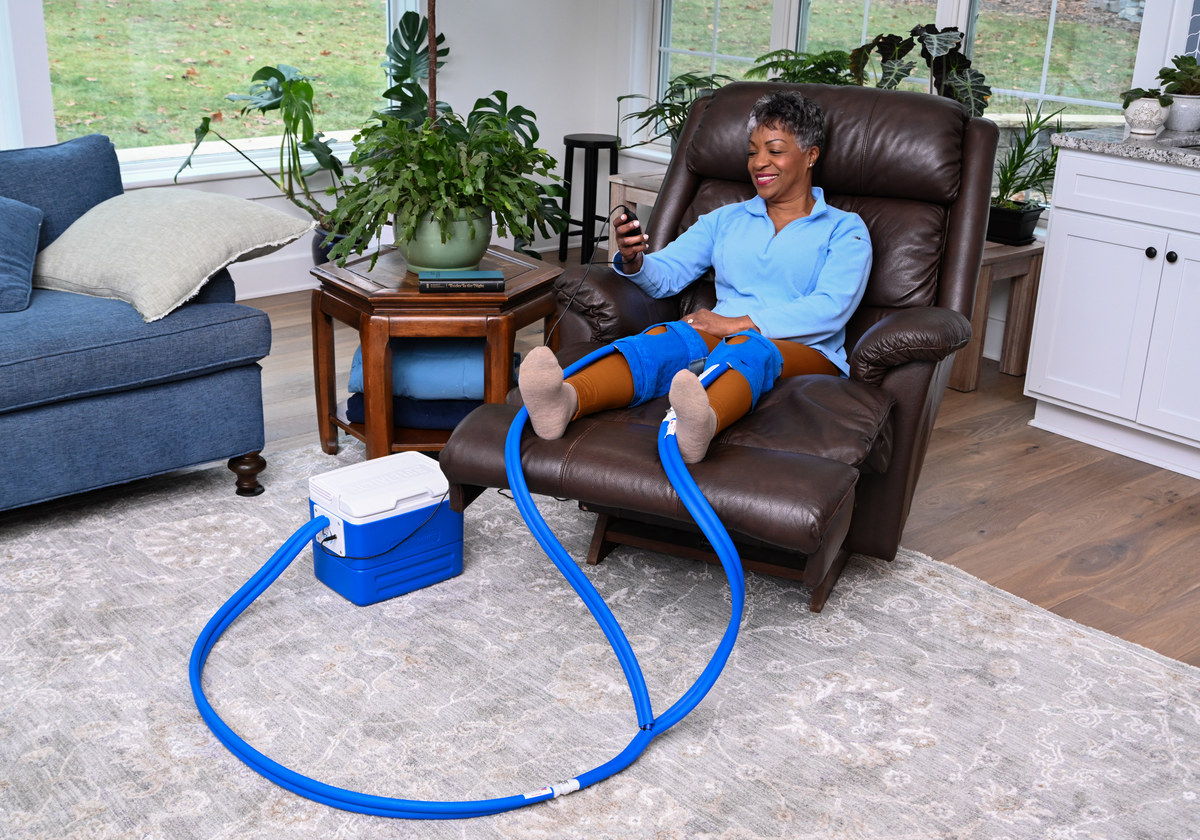 Two people sitting in a living room both using an Active Ice® Knee & Joint cold therapy system in a 16-quart cooling reservoir. One woman is using it on her elbow, while the other is using it on her knee. 