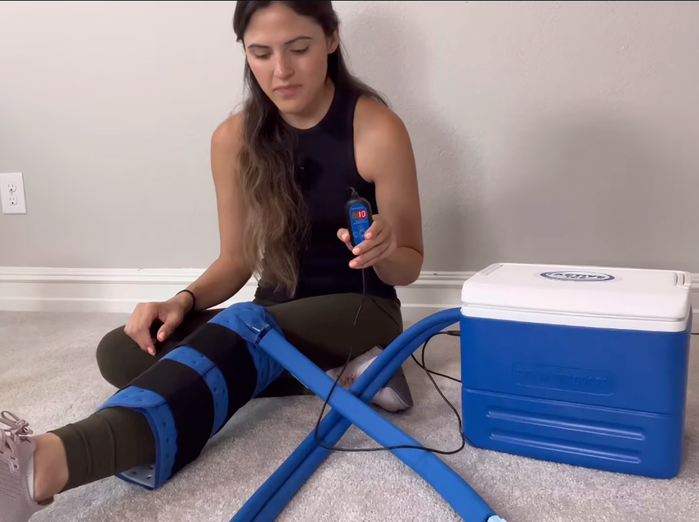 A woman sits on a carpeted floor next to a blue and white Active Ice® System using a the cooling device with tubes on her leg. She holds a small blue electronic controller.