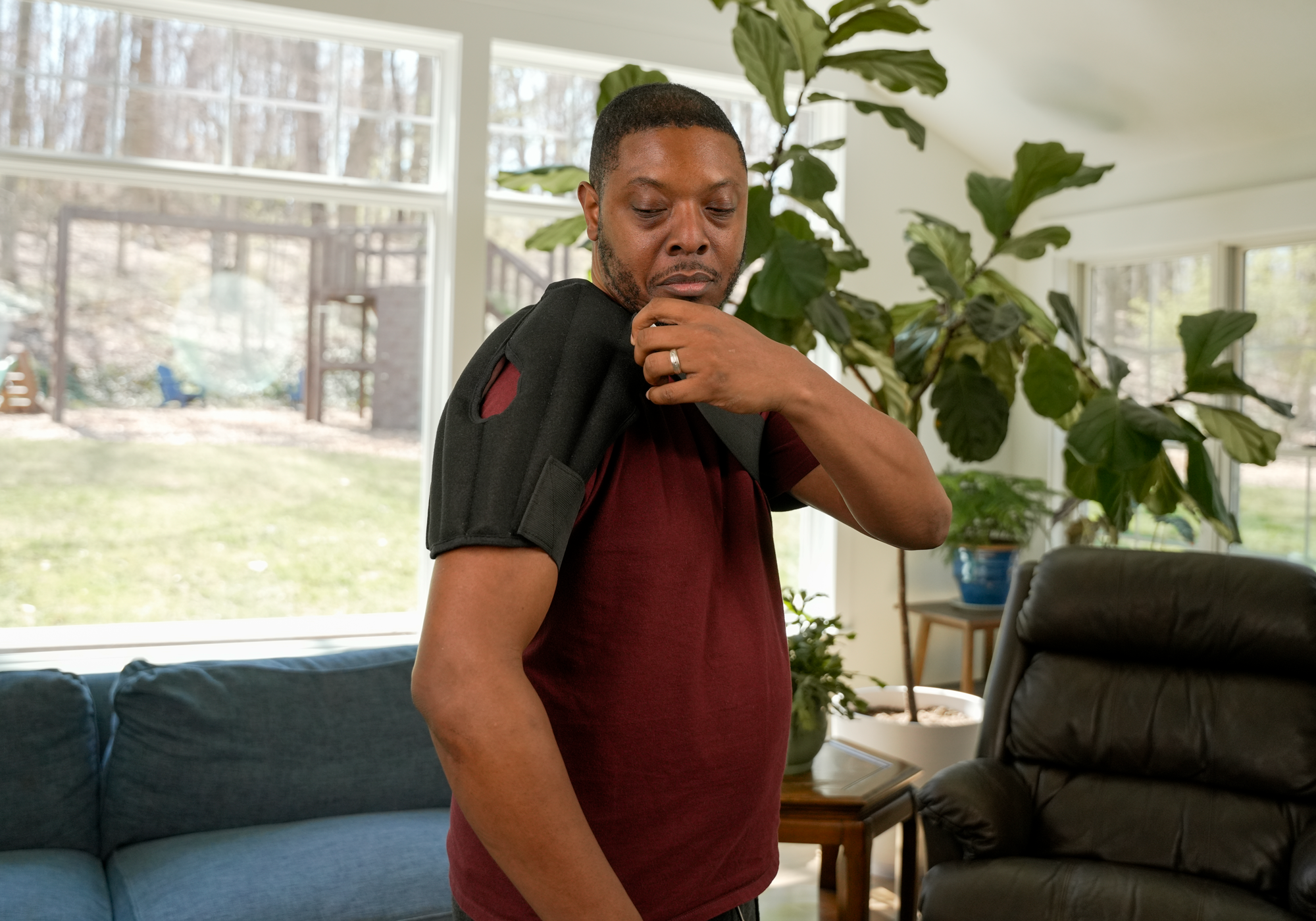 A man wearing a black moist heat wrap over a maroon t-shirt, standing in a living room with a blue couch, a large green plant, and a black leather armchair.