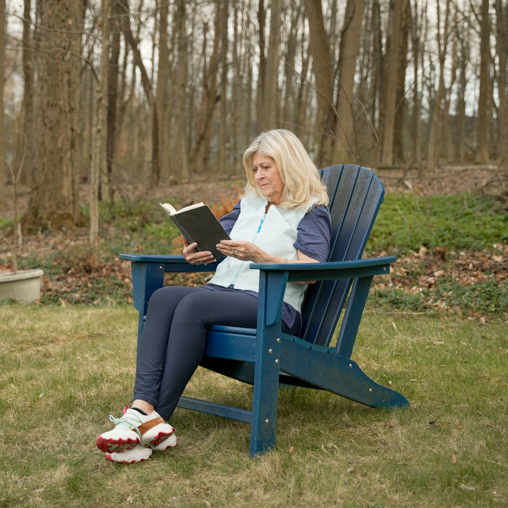 Woman reading a book in a blue Adirondack chair outdoors wearing a light blue women's fashion cooling vest.