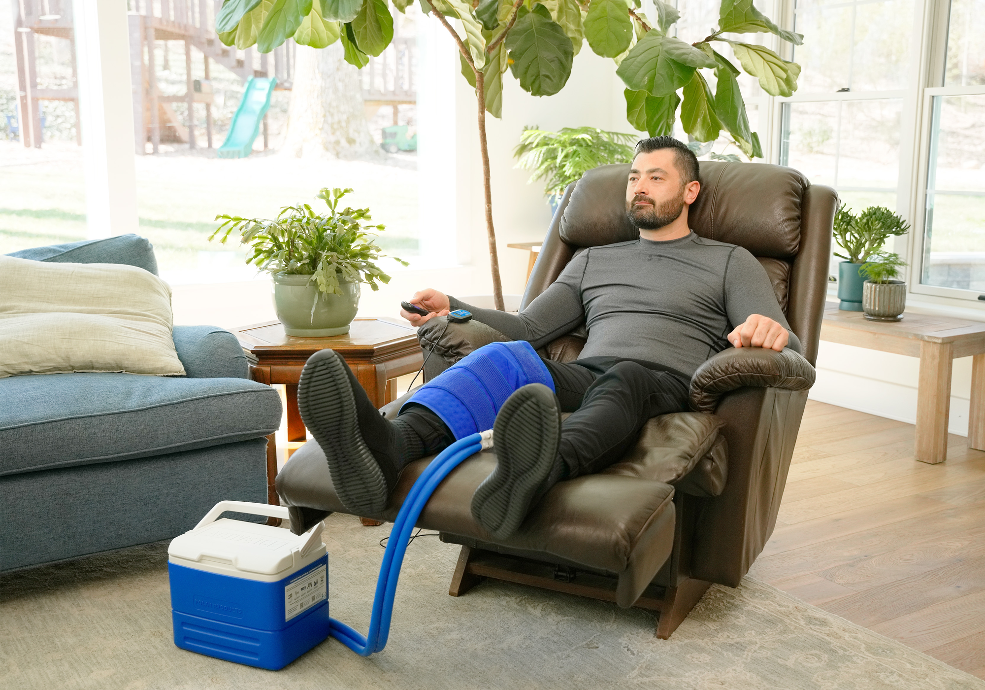 A man reclining in a brown leather armchair with his legs raised, wearing a blue full circumference knee therapy pad. A blue and white Active Ice® Cold Therapy System is connected to his leg with a blue hose, placed on the floor in a bright living room.