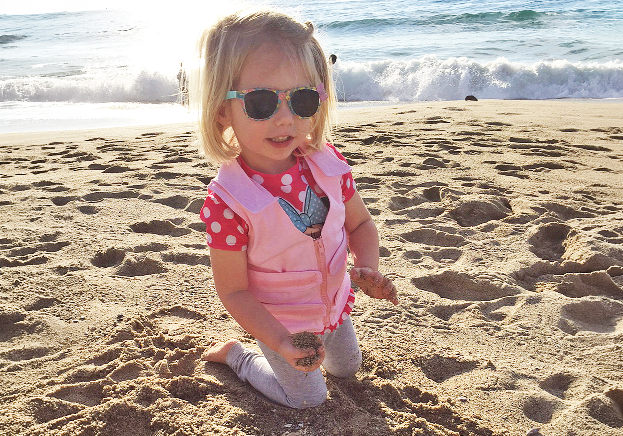 Child on a beach wearing sunglasses and a pink adjustable cooling vest while holding sand.