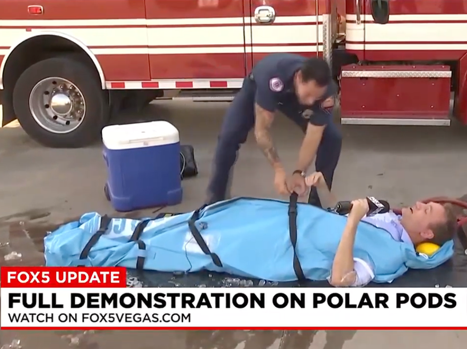 A firefighter demonstrates the use of a blue Polar Life Pod® cold water immersion system with straps on a person lying on it, with a red fire truck and cooler in the background. A Fox5 Vegas news banner is visible at the bottom.