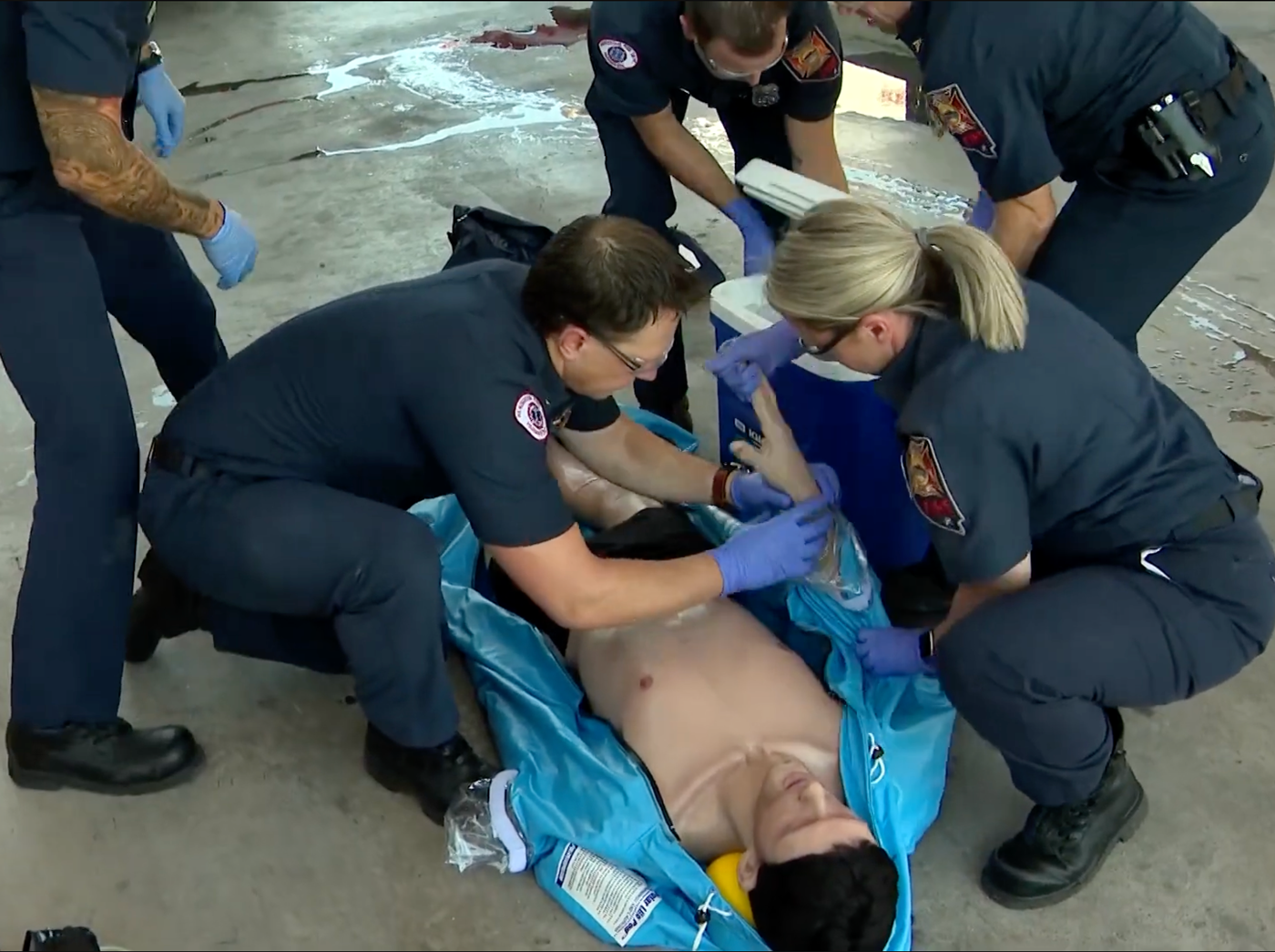 Emergency responders in dark blue uniforms and purple gloves attend to a person lying in a blue Polar Life Pod® cold water immersion system on a concrete floor.