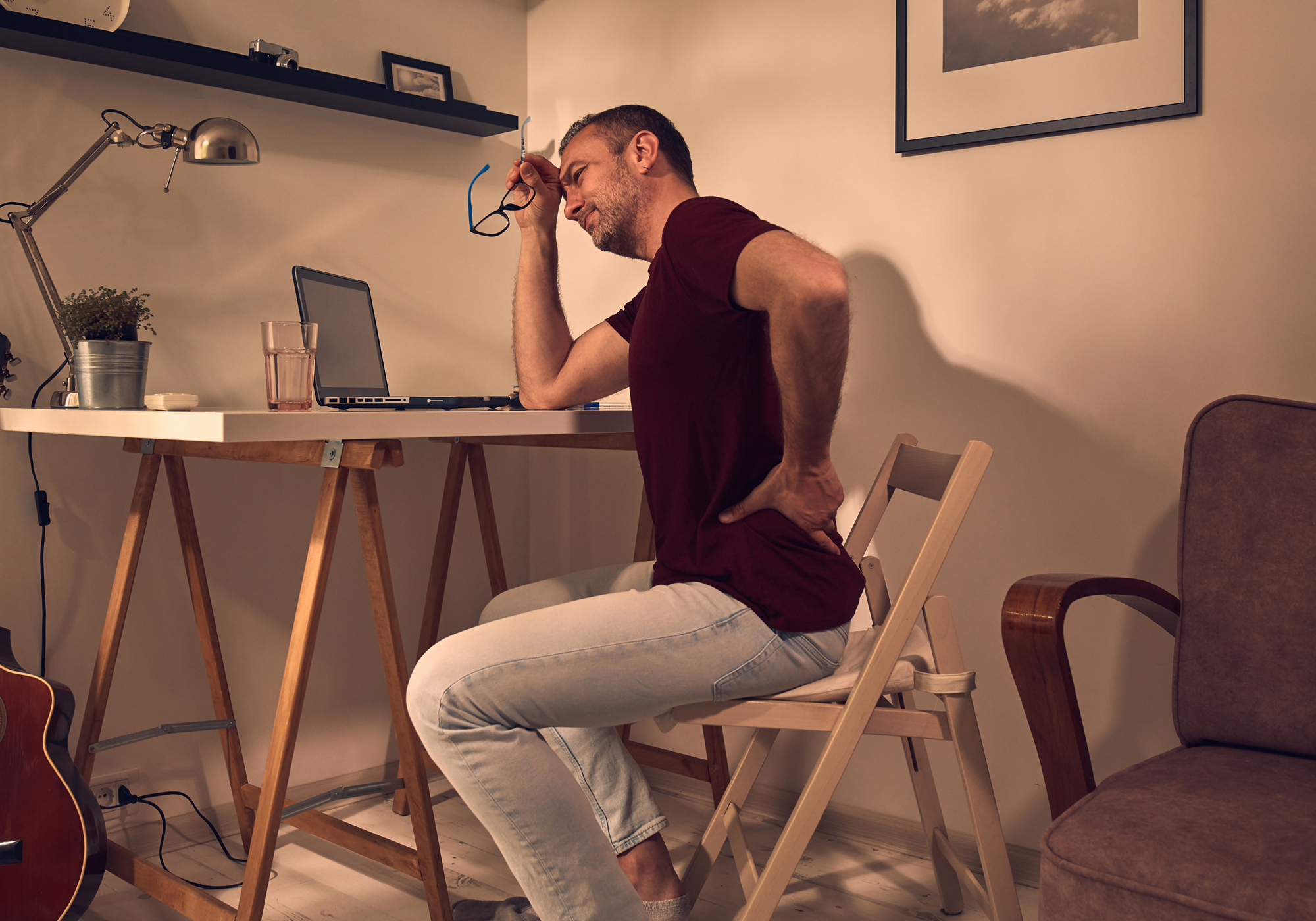 Man sitting at a desk with a laptop, holding his head and back in discomfort.