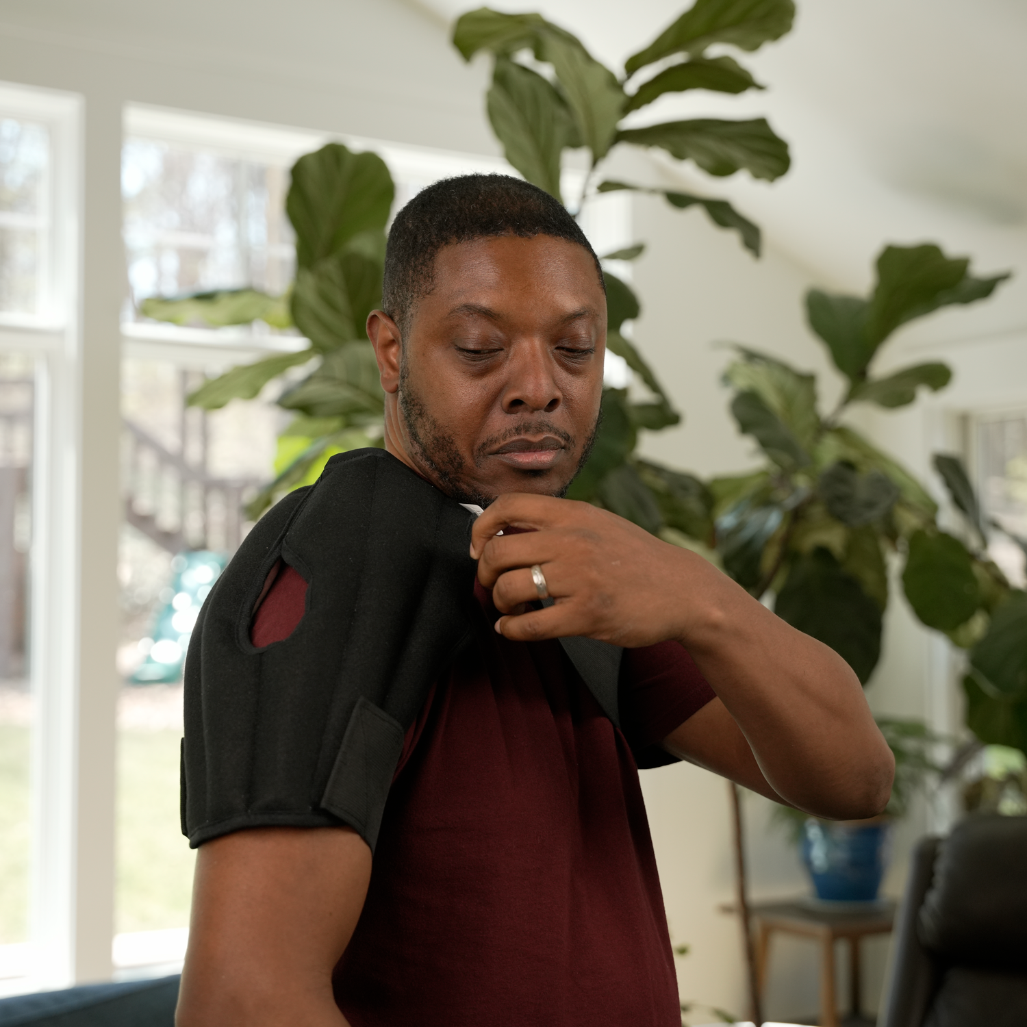 A man wearing a maroon t-shirt placing a black moist heat shoulder wrap, holding it up over his shoulder in a bright, modern room with green plants in the background.