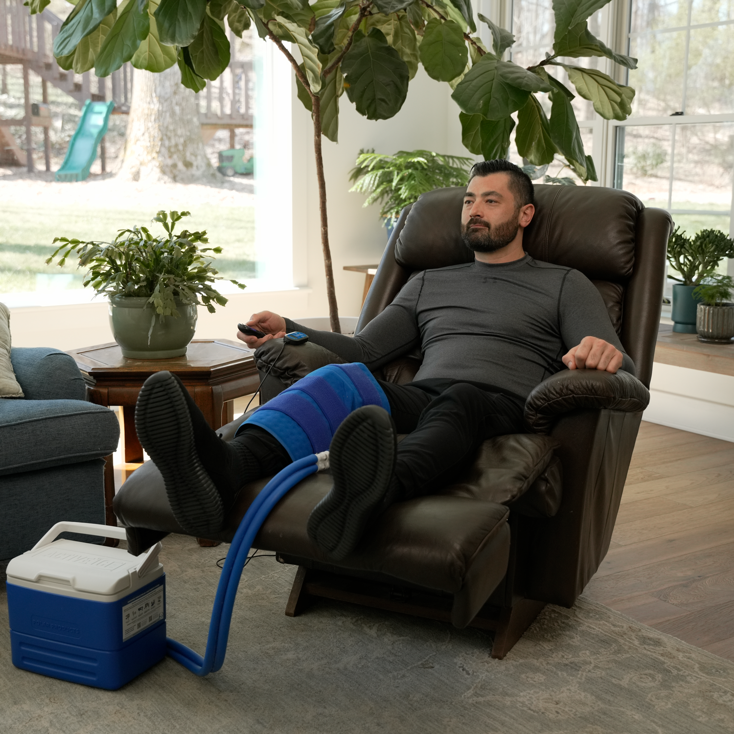 A man reclining in a brown leather armchair with his legs extended, wearing a blue full circumference knee therapy pad connected by a blue hose to a blue and white Active Ice® unit on the floor. The setting is a bright, modern living room with large windows, plants, and a tree in the background.