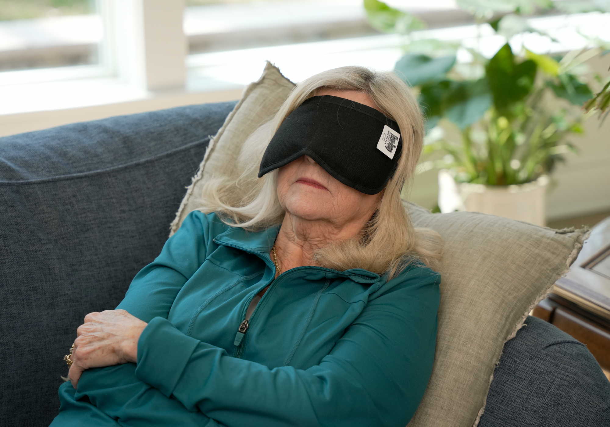 Woman wearing a black Thera-Temp® moist heat therapy mask on her eyes sitting on a couch with plants in the background