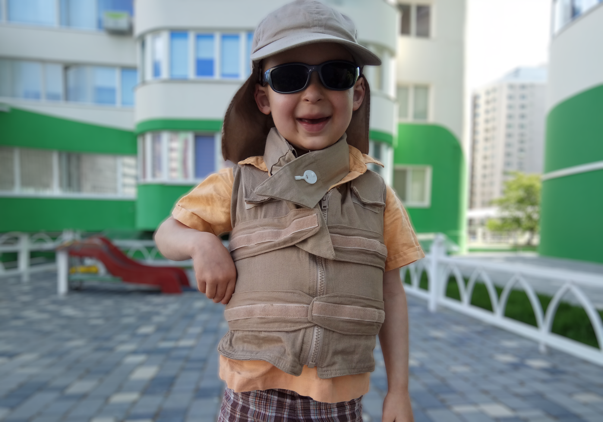 Child wearing a beige adjustable cooling vest, deluxe cooling neck wrap, sunglasses, and cap in an outdoor setting with buildings in the background.