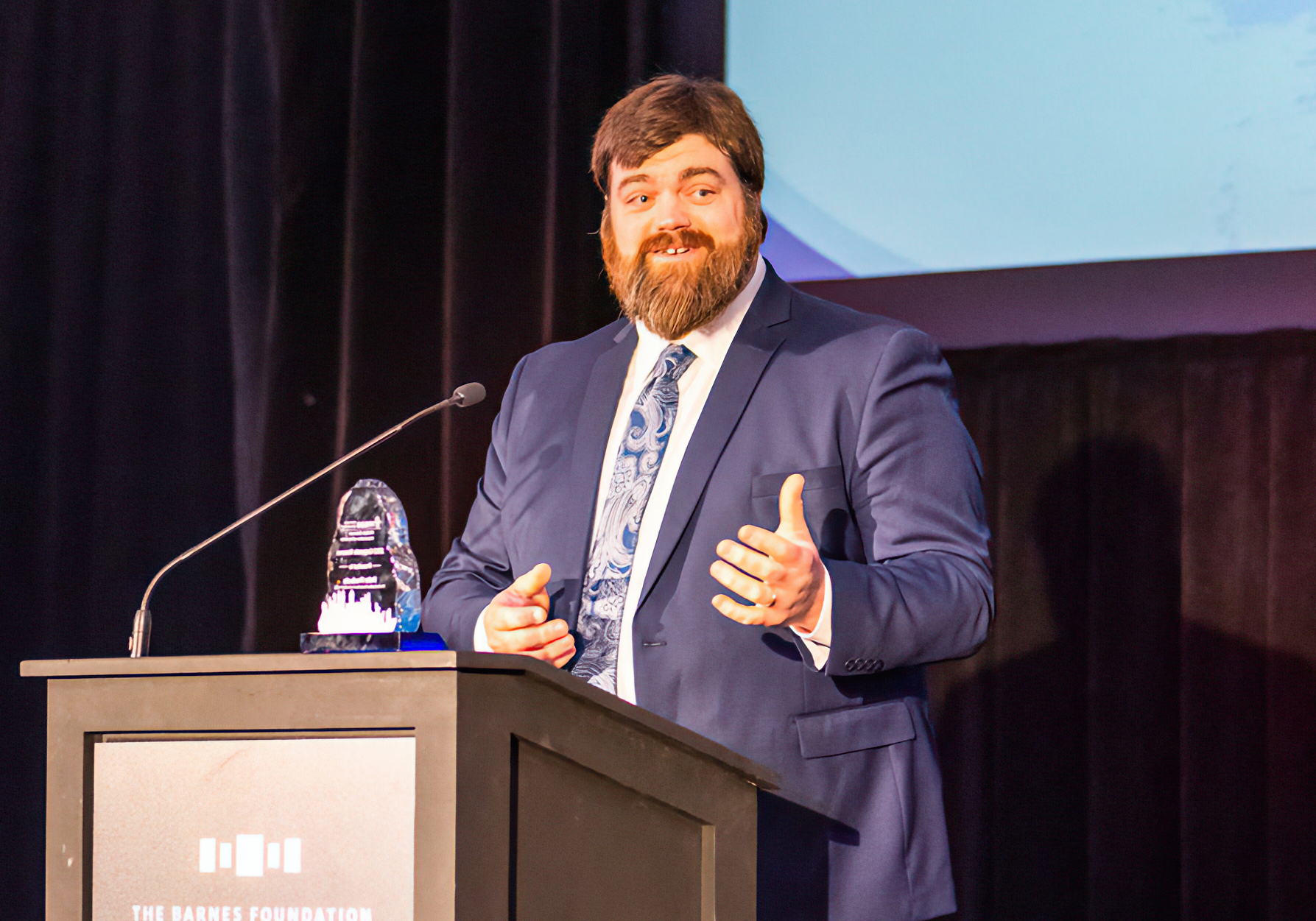 A bearded man in a navy blue suit, white shirt, and patterned tie stands behind a podium with a microphone and a clear glass award trophy on stage, with a dark curtain and screen in the background.