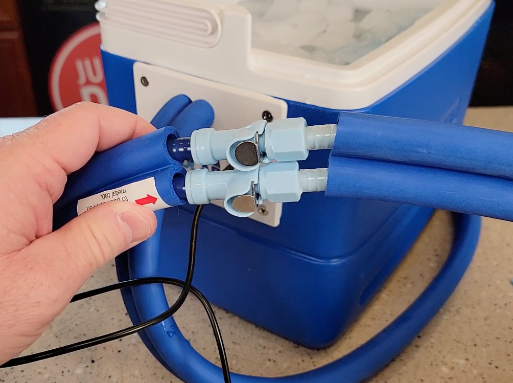A close-up of a hand holding a blue insulated tube connected to a dual T-fitting and a blue Active Ice® System, with more blue tubing looping around the cooler on a countertop.