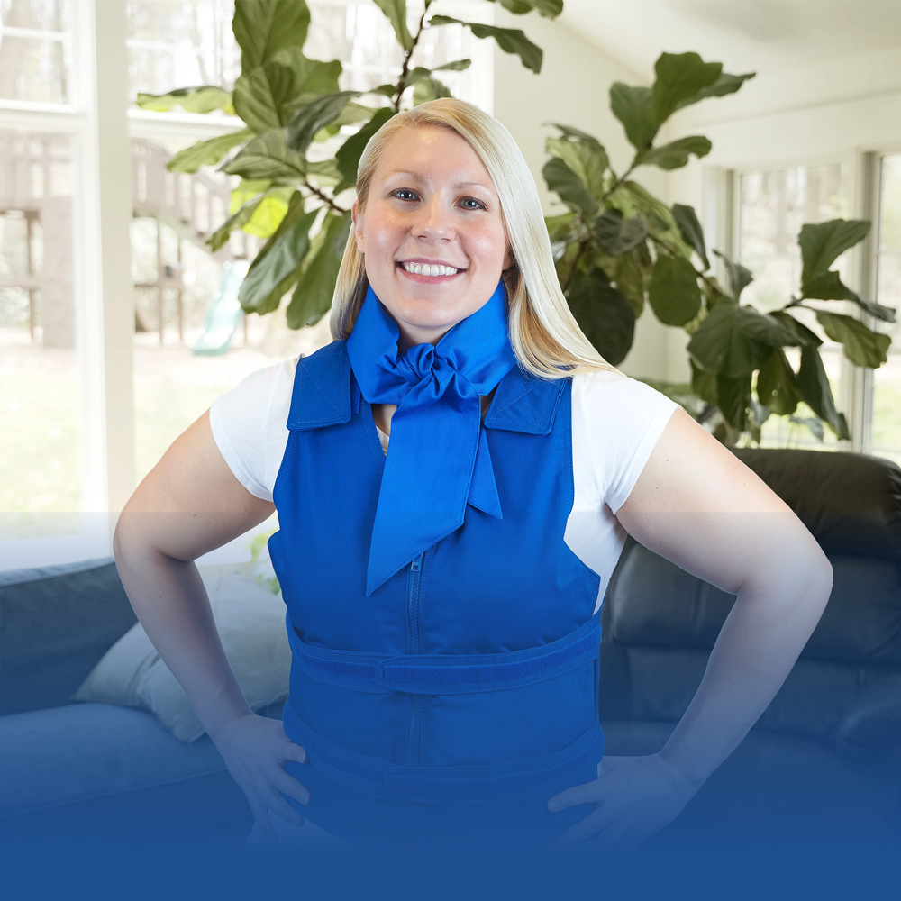 A smiling blonde woman stands indoors with her hands on her hips, wearing a bright royal blue adjustable cooling vest over a white T-shirt. Around her neck is a deluxe cooling neck wrap in blue.