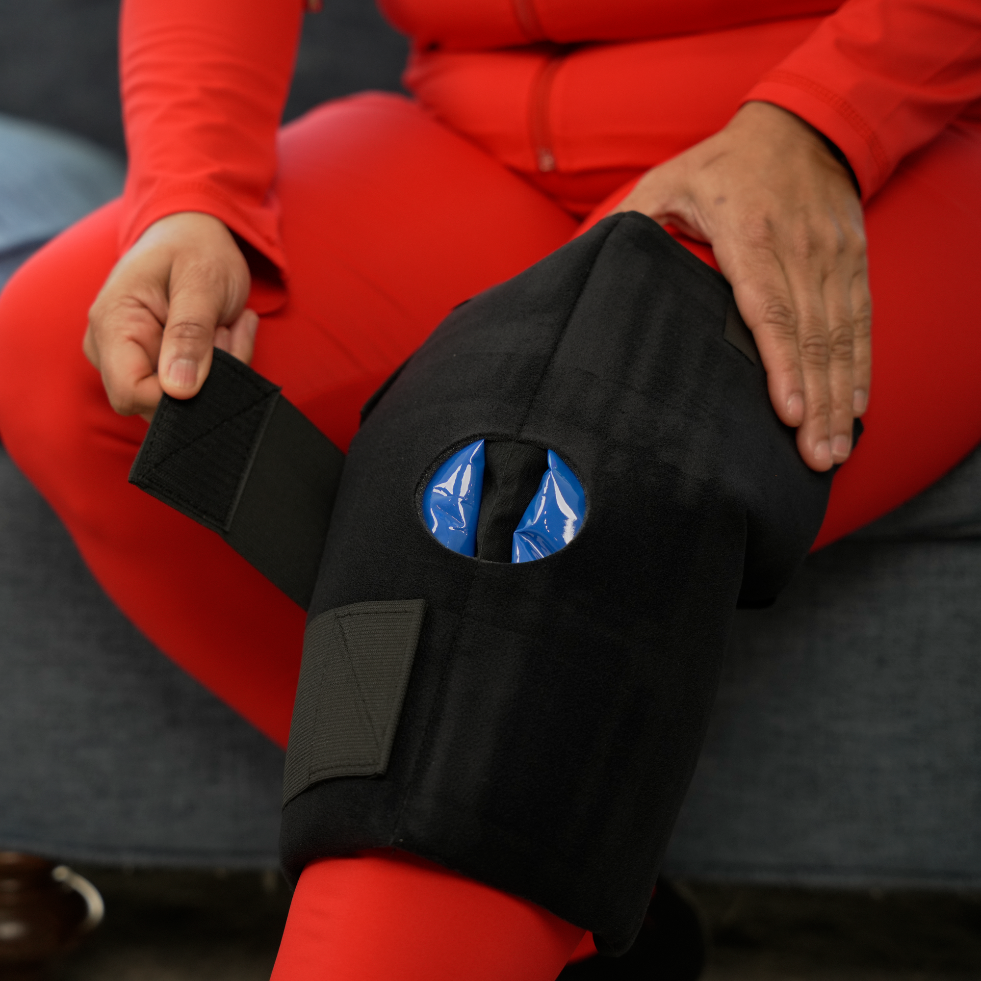 A person wearing a bright red outfit sits on a gray couch while demonstrating a black Soft Ice® knee wrap with a circular opening showing the blue Soft Ice® packs inside.