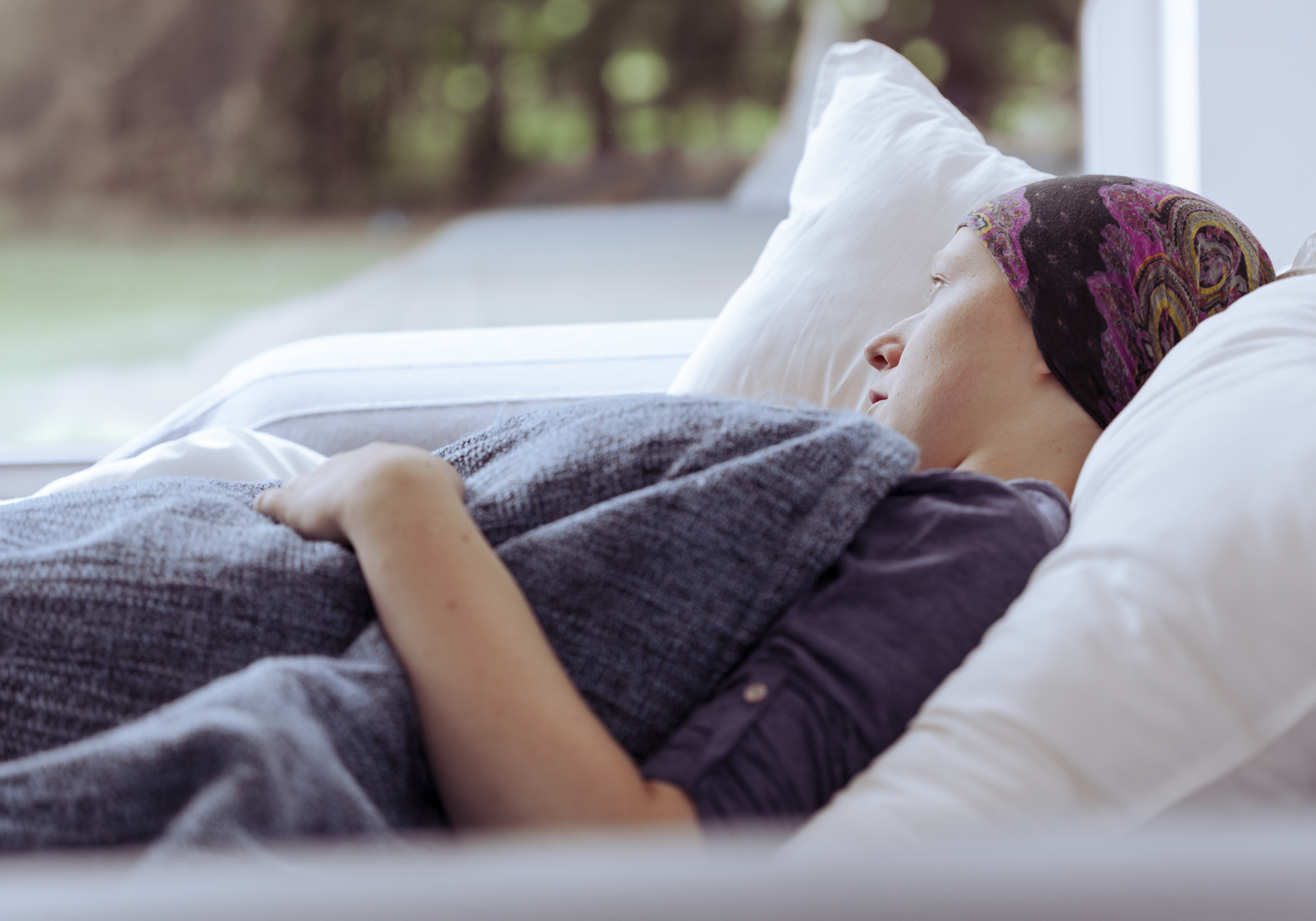 Person lying on a couch with a headscarf, possibly after chemotherapy, with a blurred outdoor background.