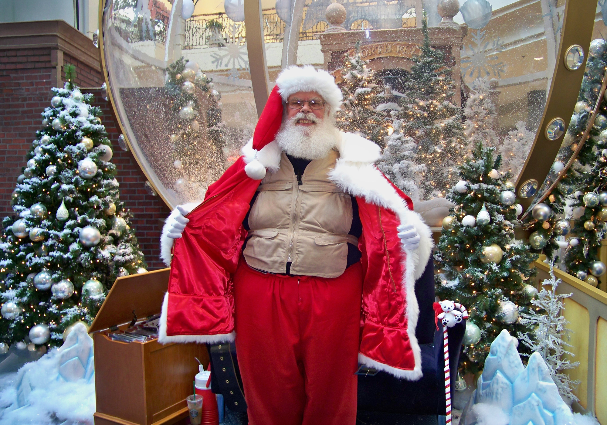 Person dressed as Santa Claus standing in a festive setting with Christmas trees and decorations.