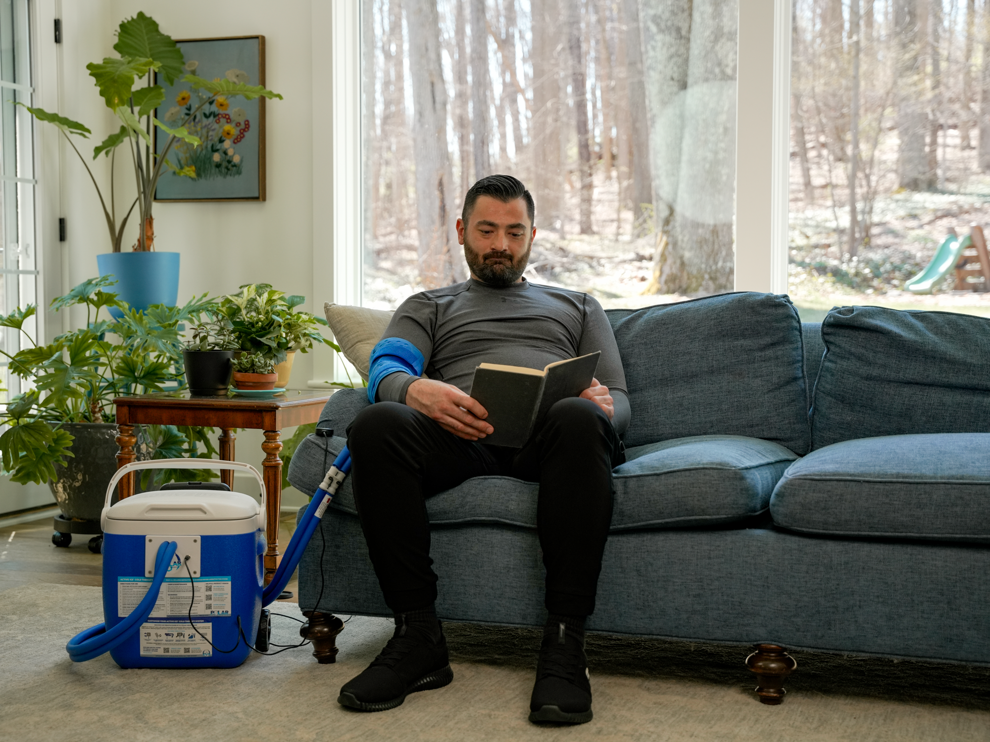 Man sitting on a couch reading a book in a living room with plants and a cooler.