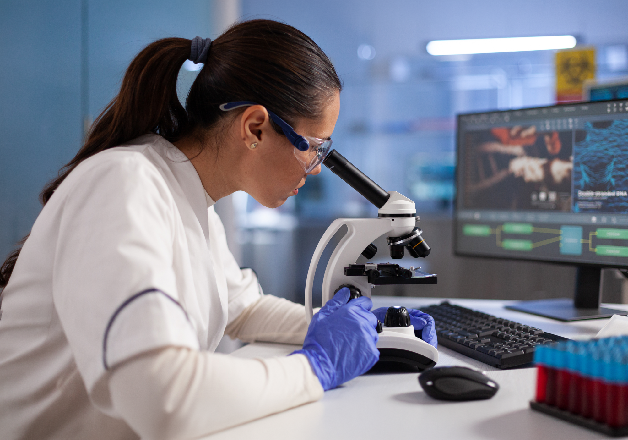 Scientist using a microscope in a laboratory setting with computer monitors in the background.