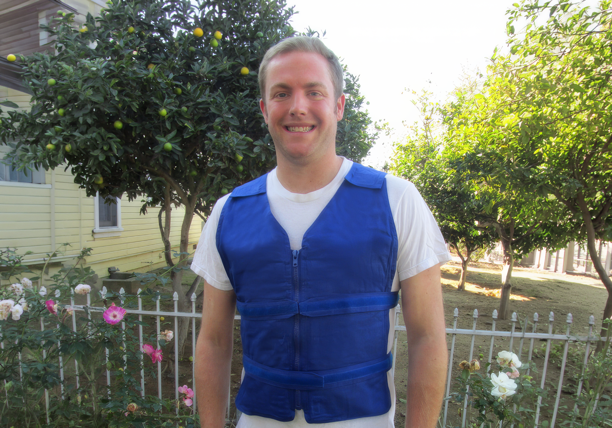Man wearing a blue adjustable cooling vest standing in front of a house with trees and flowers.