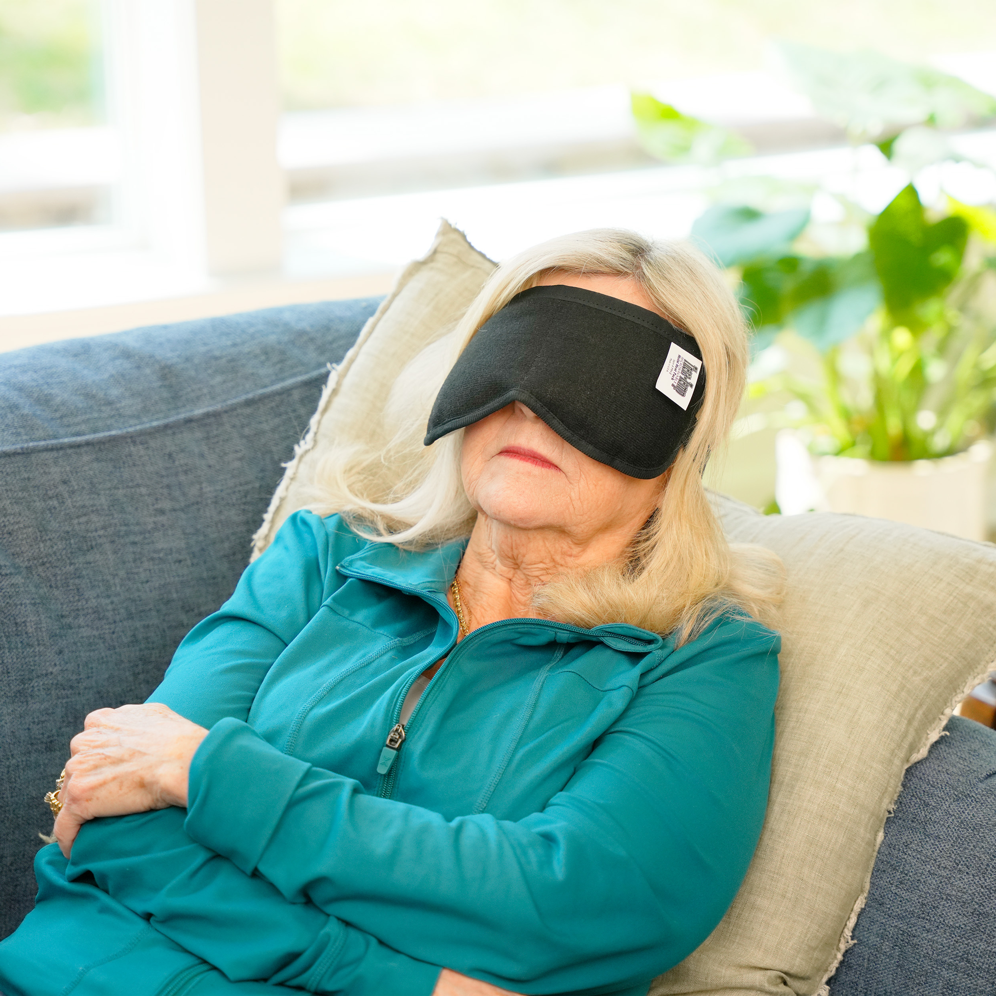 A woman reclining on a blue couch with pillows, wearing a black moist heat eye mask and a teal zip-up top. A small white label is visible on the side of the mask.