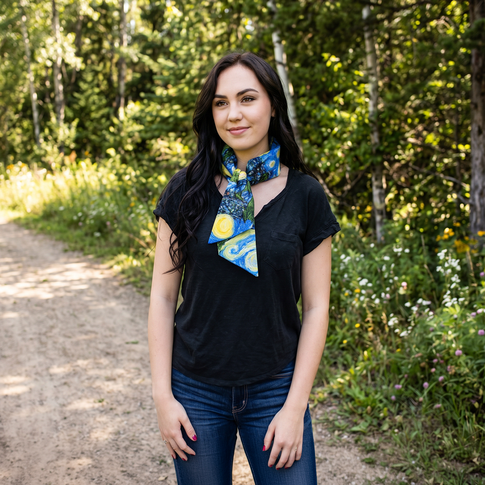 Woman standing on a forest path wearing a black top and blue jeans with a colorful cooling neck scarf.