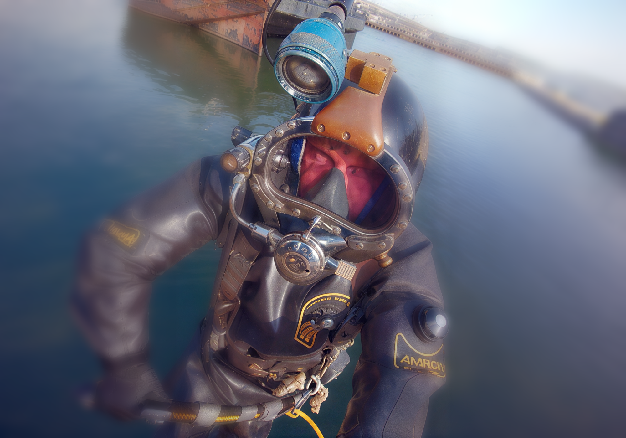 A person wearing a vintage black diving suit with a large clear helmet and brass-toned lighting equipment, standing in shallow blue water near a boat. Underneath the suit is a custom circulating cooling system provided by Polar Products. 