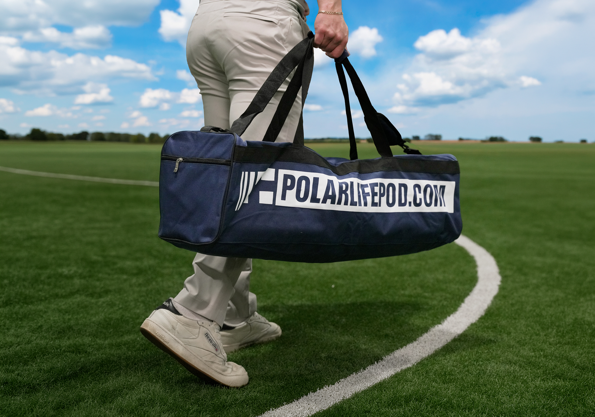A navy blue duffel bag with 'POLARLIFEPOD.COM' printed in white, carried by a person wearing khaki pants and white sneakers on a green grass field under a blue sky.