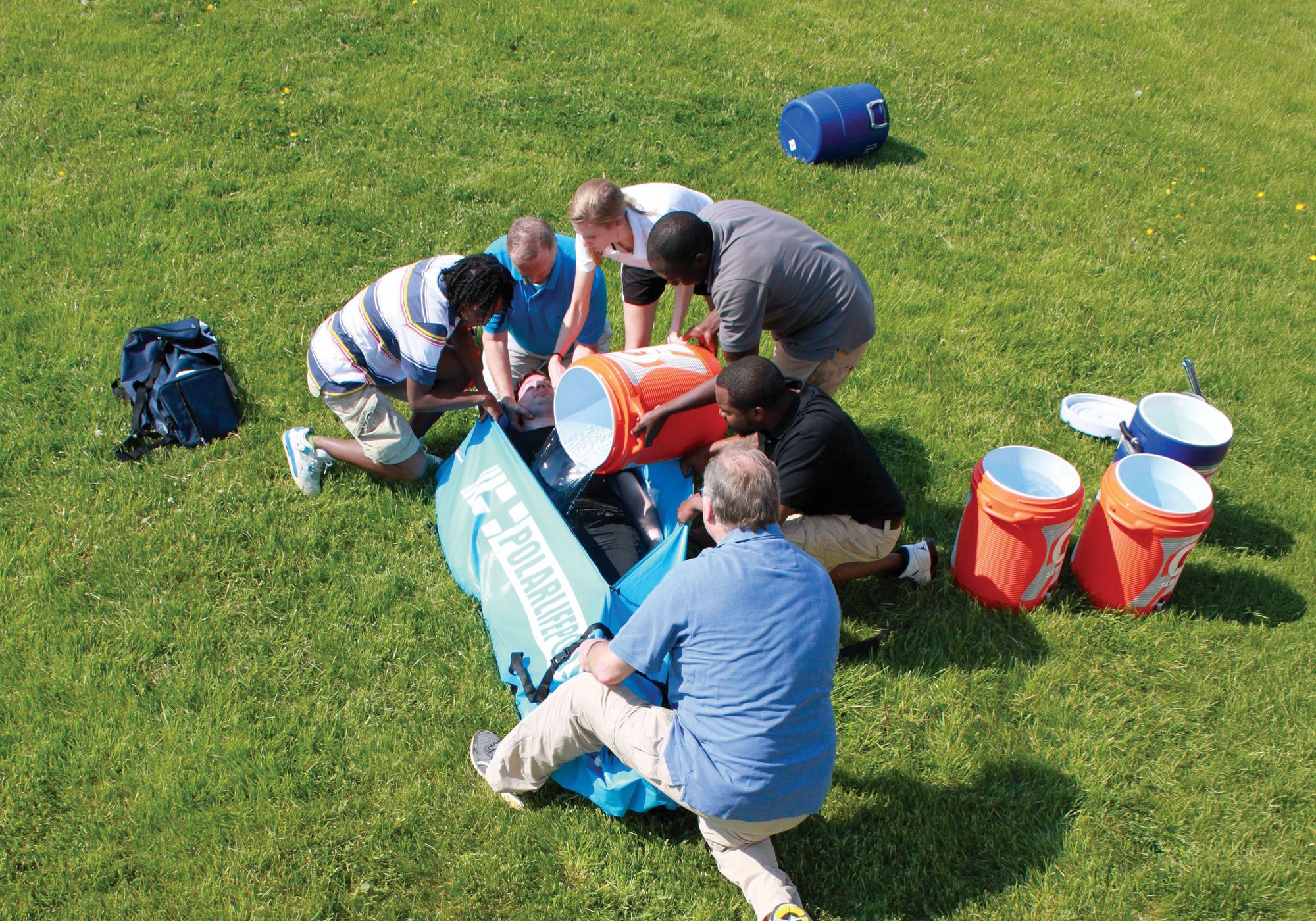 Several people sitting and kneeling on a grassy lawn are gathered around a large blue bodybag like device but its the Polar Life Pod® a cold water immersion system, with orange and blue buckets nearby.