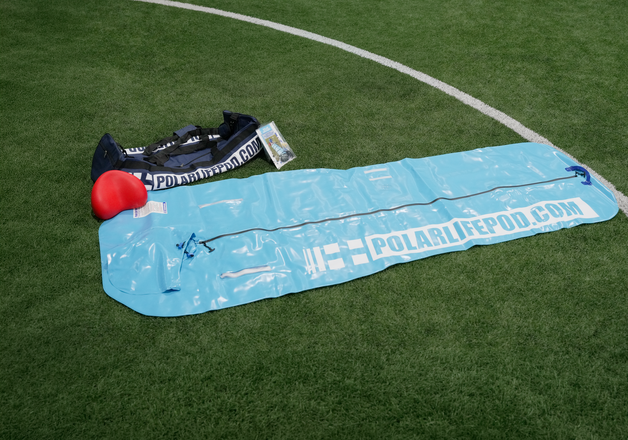 A light blue bodybag like device with white POLARLIFEPOD.COM branding is laid out on green turf next to a black carrying bag and a red floating head support pillow.