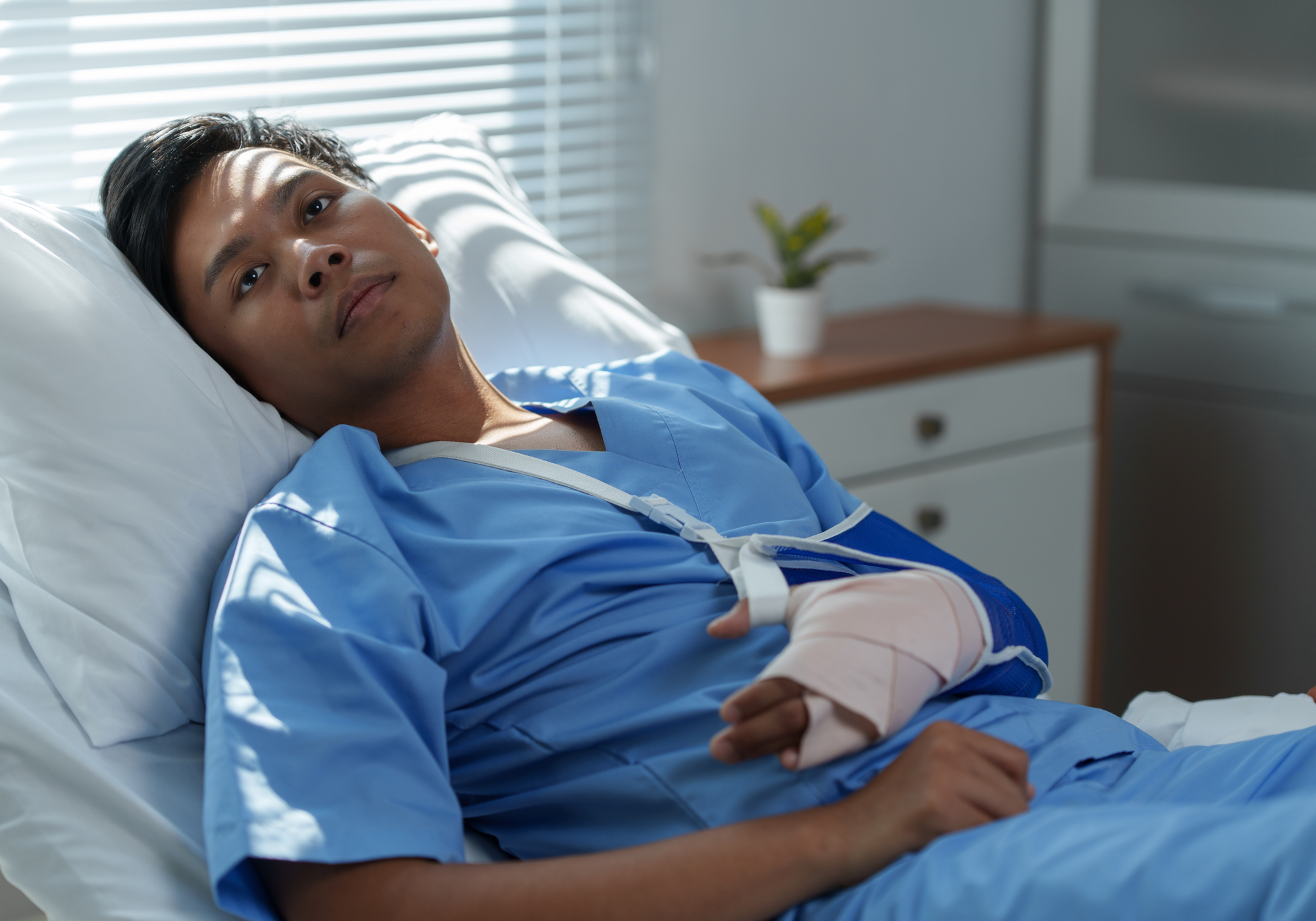 Person in a hospital bed with a bandaged arm, wearing a blue hospital gown.