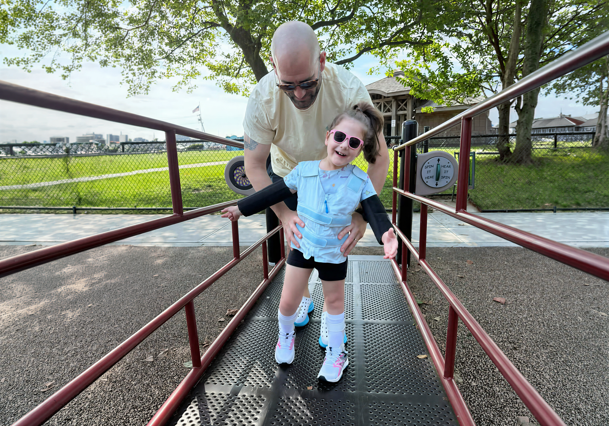 Man helping a child with blue cooling adjustable vest walk on a playground ramp with trees and playground equipment in the background.