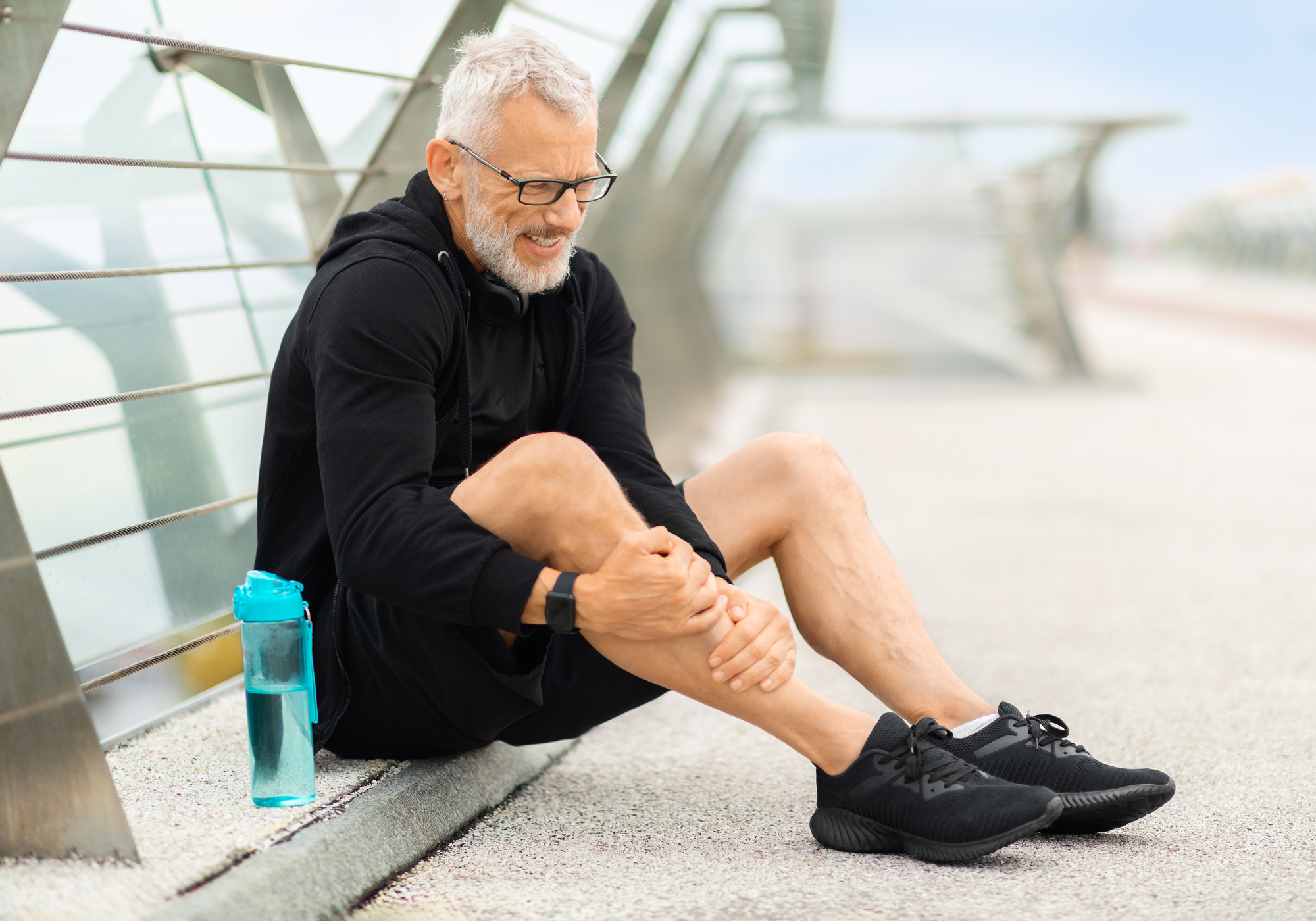 Man sitting on a bridge holding his shin, wearing black athletic wear and glasses.
