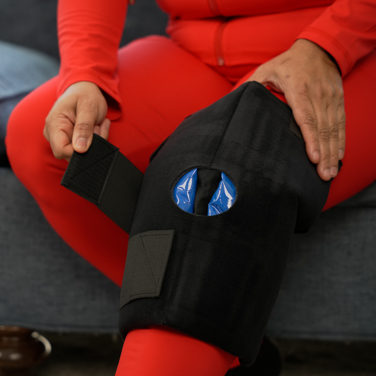 A person wearing a bright red outfit sits on a gray couch while demonstrating a black knee soft ice® wrap with blue soft ice packs.