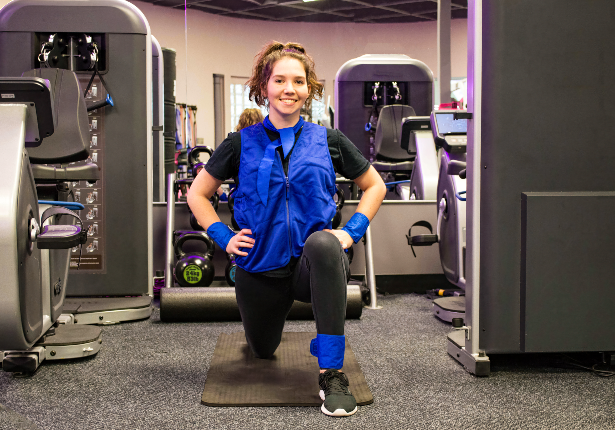 Person exercising in a gym setting wearing a blue vest and black leggings.