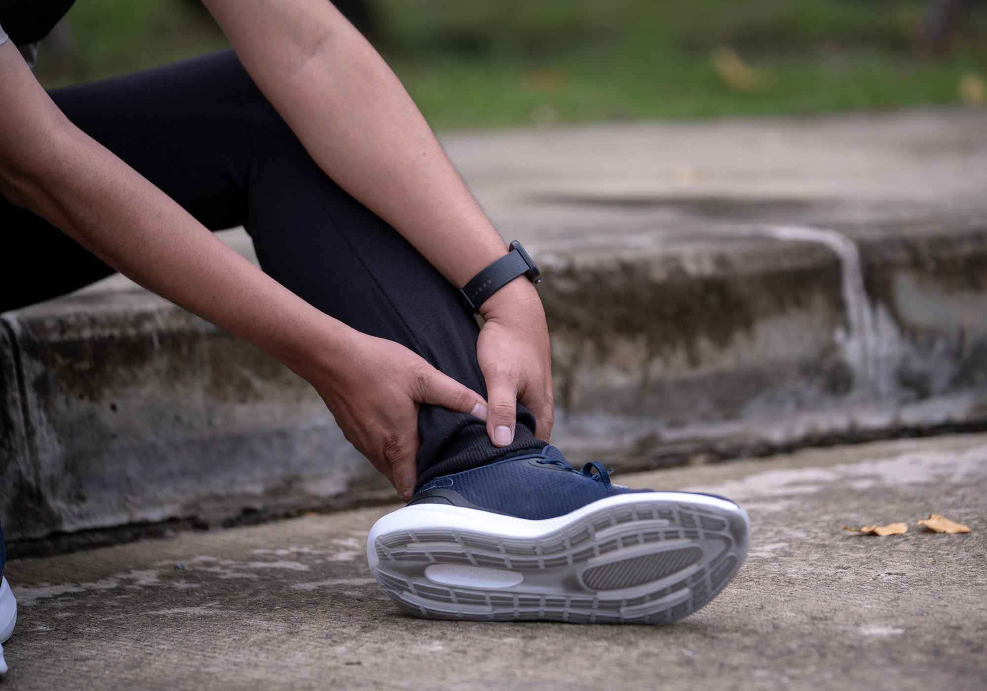 Person holding their ankle on a concrete surface with grass in the background