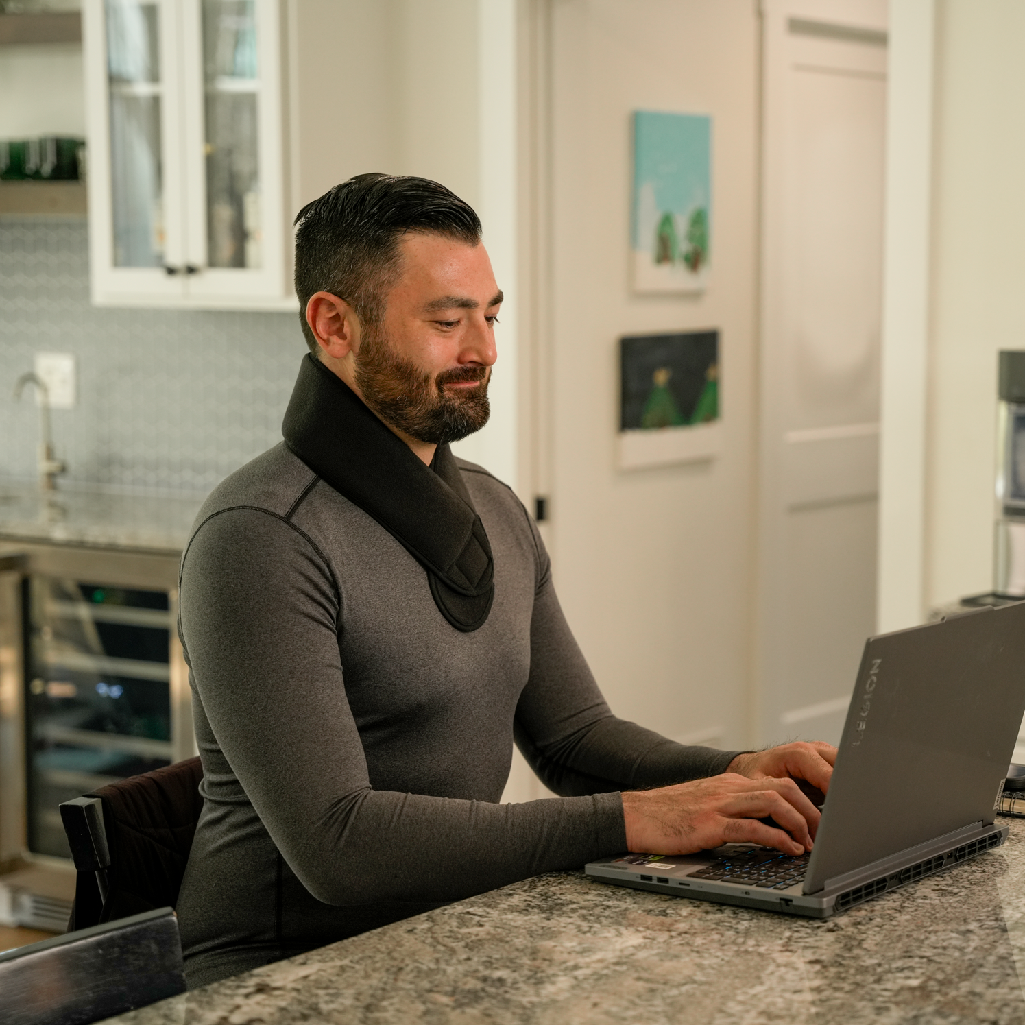 A man wearing a dark gray long-sleeve compression-style shirt with a black soft ice® neck wrap sits at a kitchen counter typing on a laptop.