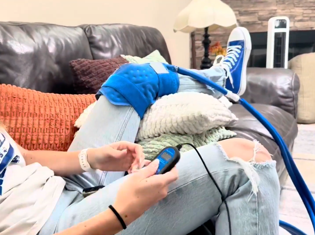 A person reclining on a dark leather couch with one leg raised, wearing a bright blue Active Ice® Therapy Pad on the raised leg. The person is holding a small blue electronic device connected by tubing, with ripped light-wash jeans and blue sneakers visible in a living room setting.