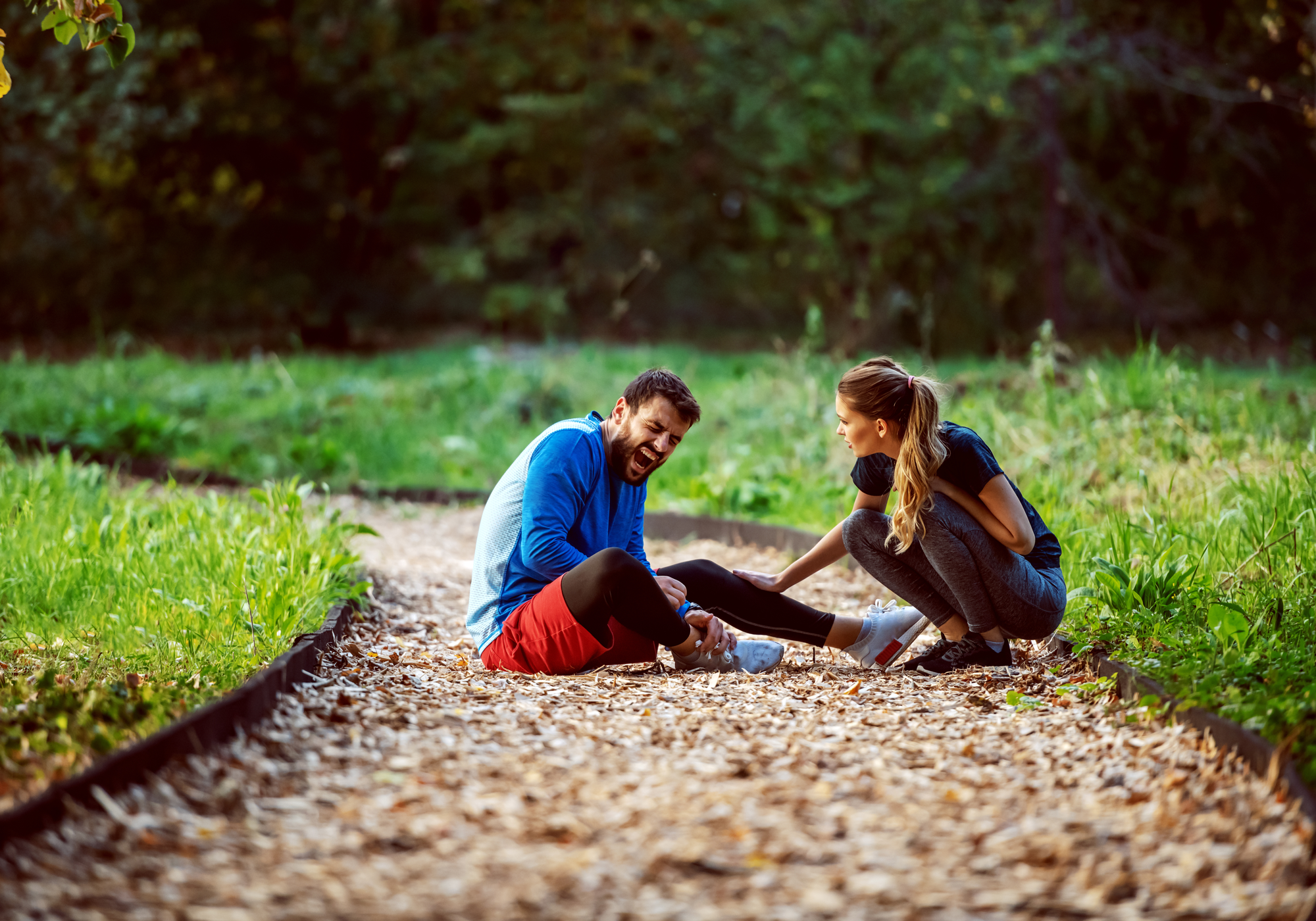 Two people sitting on a path in a park, while the man on the left is holding onto his ankle in pain.