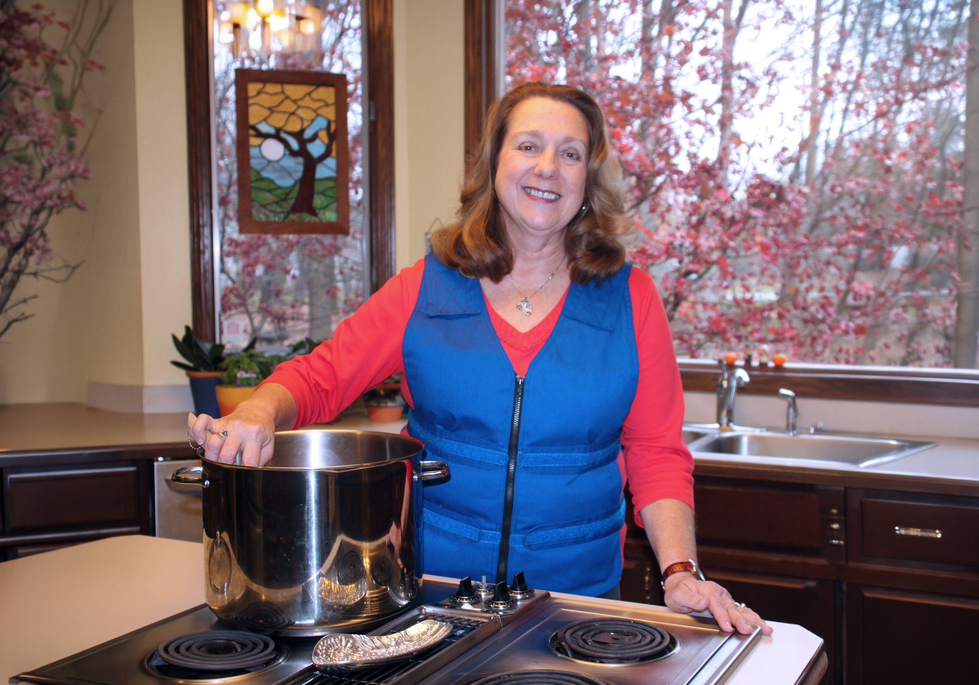 Woman standing in a kitchen with a pot on the stove, smiling at the camera.